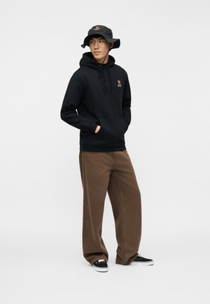 Ung mann iført svart hettegenser med rosebroderi, svart bøttehatt, brune vide bukser og svarte joggesko, stående med hendene i lommene.