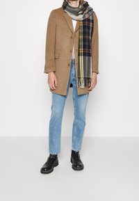 Henkilö, joka on pukeutunut kamelinväriseen takkiin, ruudulliseen huiviin, valkoiseen paitaan, vaaleansinisiin farkkuihin ja mustiin nahkasaappaisiin, seisoo valkoista taustaa vasten.