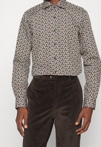 Overhemd met knopen en een wervelend patroon in bruin, goud en crème op een donkere achtergrond; gecombineerd met donkere ribfluwelen broek.