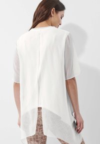 Witte doorschijnende blouse met korte mouwen, ronde halslijn en een langere achterkant. Heeft een subtiele splitdetail aan de achterzijde.