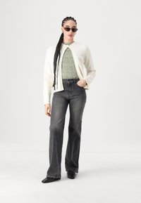 Cardigã creme, blusa verde texturizada, jeans pretos de cintura alta e sapatos pretos de bico. Acessórios incluem óculos de sol e brincos de argola.