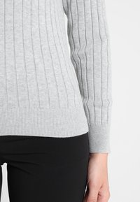 Maglione grigio a costine con maniche lunghe, caratterizzato da una vita aderente e un tessuto testurizzato. Abbinato a capi neri.