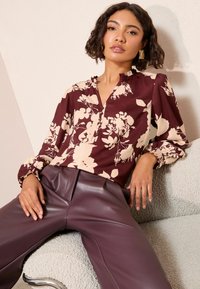 Burgundská kvetinová blúzka s dlhými rukávmi a výstrihom v tvare kľúča, skombinovaná s priliehavými, lesklými slivkovými nohavicami. Textúrované krémové pozadie.