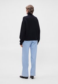 Μαύρος πλεκτός σκούφος με μανίκια και λαιμό ψεύτικου τύπου, συνδυασμένος με ανοιχτόχρωμα τζιν παντελόνια σε φαρδιά γραμμή. Η υφή φαίνεται βαριά, δημιουργώντας μια δομημένη σιλουέτα.