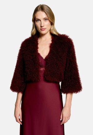 Frau mit blondem Haar, die ein burgunderfarbenes Kleid und eine passende flauschige, kurze Jacke mit Dreiviertelärmeln vor einem einfarbigen Hintergrund trägt.