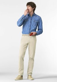 Jonge man in een spijkerhemd en beige broek die cherrytomaten van een klein rankje in zijn hand eet tegen een effen achtergrond.