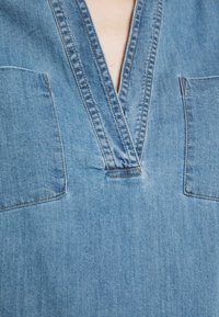 Top in denim azzurro con scollatura a V, dotato di due tasche anteriori a toppa e dettagli di cucitura visibili. La texture è liscia e leggermente usurata.