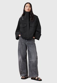Giacca nera oversize con due grandi tasche frontali, colletto corto e zip. Abbinata a jeans grigi strappati a gamba larga e sandali neri.