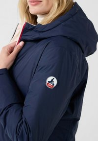Giubbotto imbottito blu navy con cappuccio, dotato di una cerniera rossa e di una patch con il logo JOTT sulla manica. Materiale morbido e leggero con una consistenza liscia.