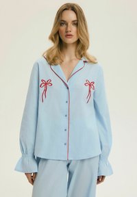 Młoda kobieta o blond włosach, ubrana w jasnoniebieską koszulę na guziki i spodnie, z czerwonymi wyszywanymi kokardkami na klatce piersiowej oraz rozszerzanymi mankietami rękawów.