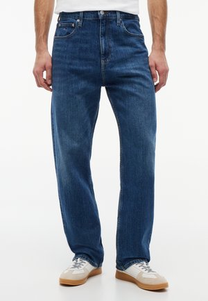 Muškarac obučen u plave traperice ravnog kroja s bijelim i bež tenisicama, stoji ispred jednobojne bijele pozadine.