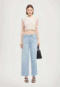 Femeie îmbrăcată cu un top cropped fără mâneci bej, blugi largi de culoare albastră deschisă, sandale negre și ținând o geantă de mână neagră.