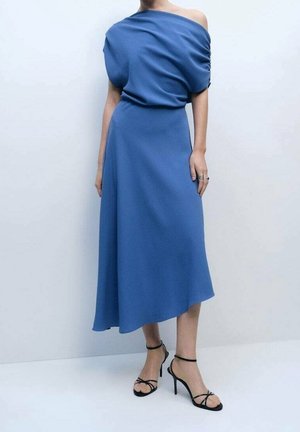 Mujer con vestido midi azul de un solo hombro con parte superior drapeada y sandalias negras de tacón con tiras, posando contra un fondo claro y liso.