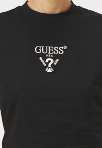 Crna majica od pamuka s rebrastim okruglim izrezom. Sadrži bijeli vezeni "GUESS" logo i tekst. Jednostavan i prilagođen dizajn.