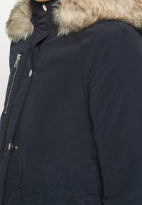 Navy blue parka jacket featuring a faux fur collar, zip front, and snap buttons, with a smooth texture and structured fit.