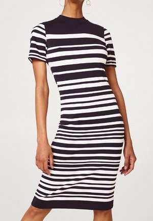 Kobieta w dopasowanej sukience do kolan z krótkim rękawem, w poziome czarno-białe paski, z kolczykami w kształcie kół.