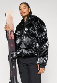 Μαύρο γυαλιστερό φουσκωτό μπουφάν με γεωμετρικό ράψιμο, φερμουάρ σε όλο το μήκος και κουκούλα. Το μοντέλο κρατάει ένα σκι με δέστρες. Το σύνολο ολοκληρώνουν σκούρα σκι παντελόνια.