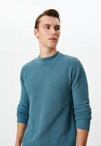 Dzianinowy sweter z okrągłym dekoltem w kolorze turkusowym, z fakturowanym wzorem i ściągaczami na mankietach. Materiał wygląda na miękki i lekki.