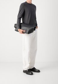 Homme portant une chemise à manches longues gris foncé, un pantalon blanc à jambes larges, des chaussures noires, tenant un grand sac en cuir noir plié sur un fond blanc.