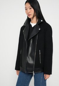 Μαύρο μπουφάν που συνδυάζει υφασμάτινη υφή και λείο τεχνητό δέρμα. Διαθέτει φαρδύ γιακά, φερμουάρ μπροστά και πλευρικές τσέπες. Σχεδιασμένο με άνετη εφαρμογή.