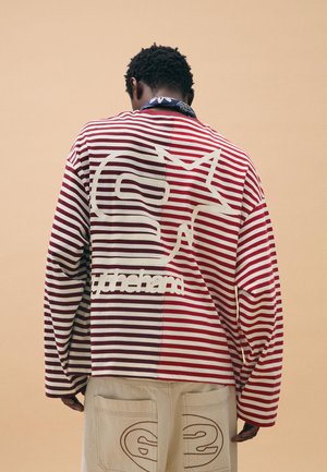 Man wearing a red and white striped long-sleeve shirt with a cat outline and text on the back, beige pants with sewn designs on pockets.