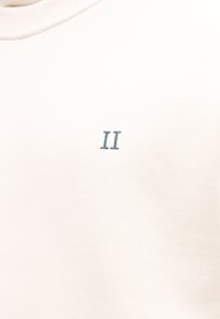 Λευκό φούτερ με απαλή υφή και μικρή μπλε κεντημένη λεπτομέρεια "II" στο μπροστινό μέρος. Απλός και μινιμαλιστικός σχεδιασμός.