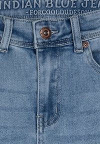 Ljusblå denimjeans med en lätt blekning, med en brun metallknapp och en sydd midjebandag med texten "INDIAN BLUE JEANS."