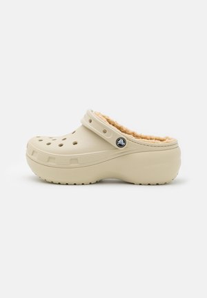Cream-colored clog made of synthetic material, featuring a soft, beige fur lining, vent holes, and a black emblem on the side.