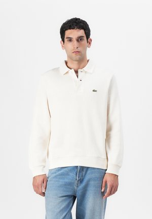 Polo crème à manches longues en tissu texturé, doté d'une patte de boutonnage à trois boutons et d'un logo brodé vert sur la poitrine gauche.