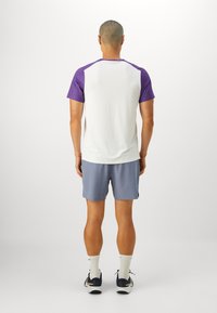 T-skjorte med korte ermer i hvit kropp og lilla raglan-ermer, kombinert med lyseblå shorts og grå sko, sett fra baksiden.