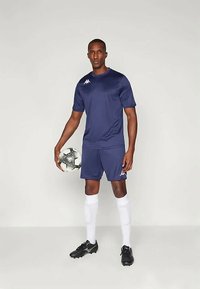 Jūras spēļu krekls un šorti, izgatavoti no gludas auduma, apvienoti ar baltām zeķēm un melniem futbola apaviem. Tur rokās melnbaltu futbolbumbu.