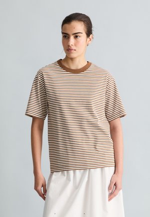 Junge Person trägt ein braun-weiß gestreiftes kurzärmeliges Shirt und einen weißen Rock, steht vor einem einfarbigen hellgrauen Hintergrund.