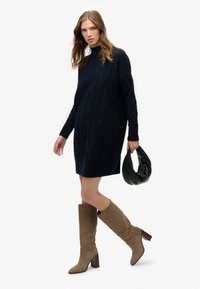 Vestido suéter de punto grueso en azul marino con mangas largas, combinado con botas beige hasta la rodilla y tacón alto. La modelo sostiene un bolso negro con textura.