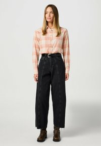 Camisa de botón de manga larga en cuadros naranja y crema, combinada con jeans de pierna ancha negros de cintura alta y botas de tobillo marrones con hebillas.