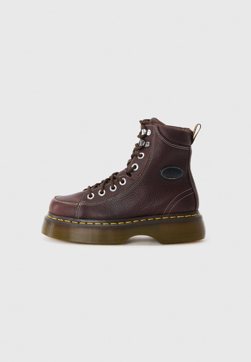 Botte montante en cuir marron avec semelle épaisse translucide, coutures jaunes, œillets métalliques et col matelassé à la cheville sur fond uni.