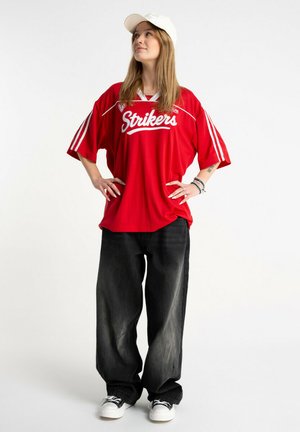 Młoda kobieta stoi z rękami na biodrach, ubrana w czerwoną koszulkę sportową "Strikers", czarne szerokie spodnie, białą czapkę i trampki.