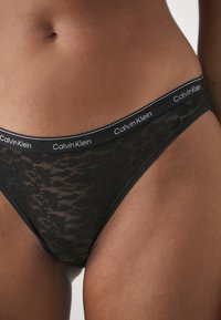 Zwarte kanten string met een gladde tailleband met het "Calvin Klein" logo. Doorschijnende textuur met geometrische patronen, die een minimalistisch ontwerp biedt.