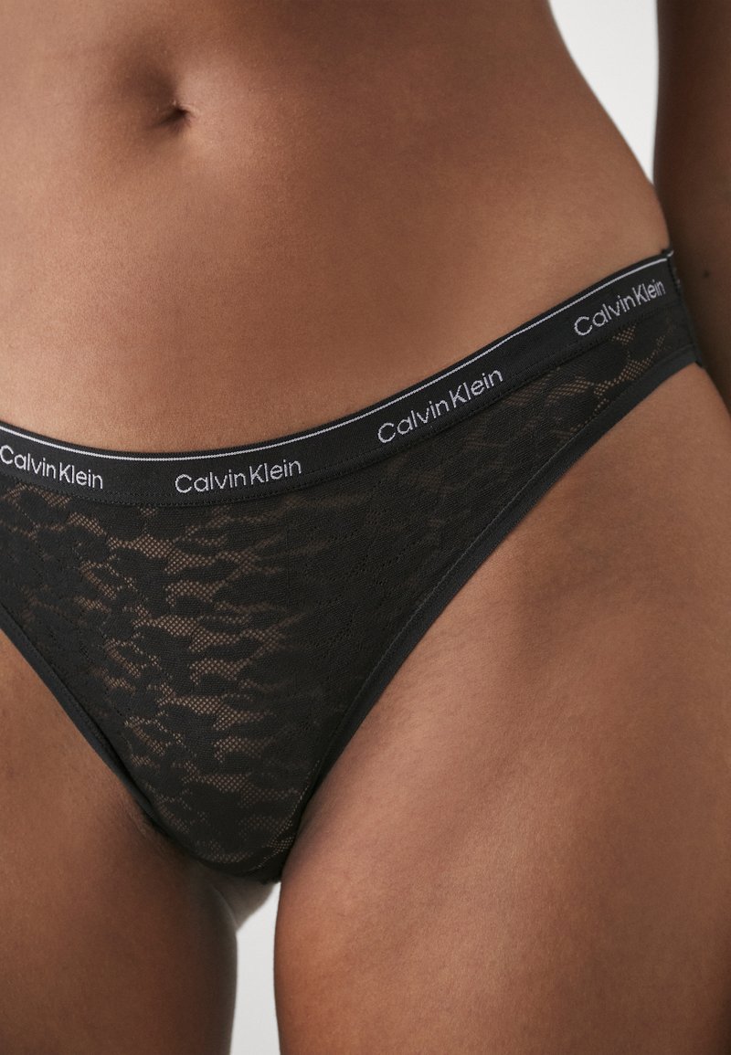 Czarny koronkowy string z gładkim paskiem w talii, na którym widnieje logo "Calvin Klein". Przezroczysta tekstura z geometrycznymi wzorami, oferująca minimalistyczny design.