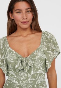 Robe verte à motif avec des manches flottantes courtes, décolleté profond et un détail à nouer au niveau de la poitrine. Tissu texturé avec des motifs de feuilles blanches.