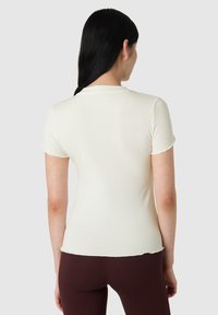 Krémový žebrovaný top s krátkým rukávem, kulatým výstřihem a vlnitým lemem, doplněný o tmavě hnědé kalhoty. Zadní pohled ukazuje úzkou siluetu.