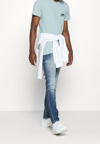 Jasnoniebieska bawełniana koszulka z małym czarnym logo, noszona pod białą bluzą zawiązaną w talii, w połączeniu z przetartymi niebieskimi dżinsami i białymi trampkami.