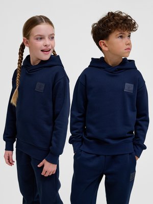 Deux enfants, une fille et un garçon, portant des sweats à capuche bleu marine assortis et des pantalons, sont debout côte à côte devant un fond uni.
