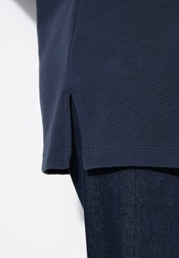 Marineblaues, strukturiertes Hemd von der Seite gezeigt, mit einem seitlichen Schlitz am Saum, kombiniert mit dunklen Jeans.