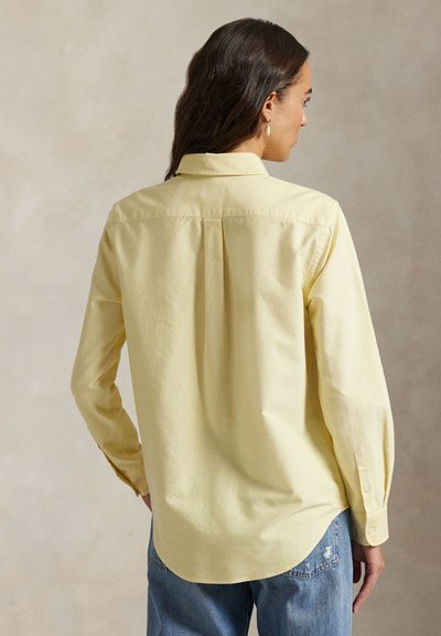 Frau mit langen dunklen Haaren, die ein blassgelbes Langarmshirt und blaue Jeans trägt, von hinten vor einem einfarbigen Hintergrund gezeigt.