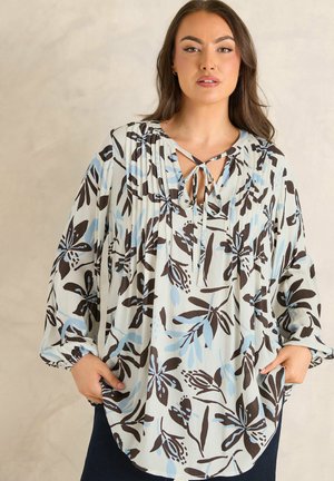 Kobieta ubrana w luźną, długorękawową bluzkę z niebiesko-brązowym kwiatowym wzorem, stojąca z rękami w kieszeniach na neutralnym tle.