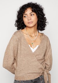 Beige omslagströja i mjukt, texturerat stickat med V-ringning och knytdetalj i midjan, lager på lager över en vit linne.