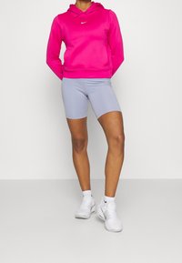 Personne portant un sweat à capuche Nike rose vif, un short de vélo violet clair, des baskets Nike blanches, debout les mains derrière le dos contre un fond uni.