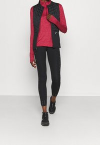 Gilet imbottito nero con zip, maglia rossa a maniche lunghe, leggings neri con dettagli bianchi decorativi e scarpe sportive nere.