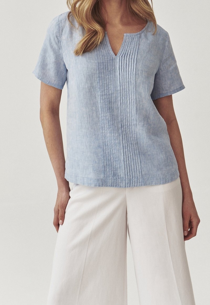 Vrouw in een lichtblauw kortemouw blouse met verticale plooien en witte wijde broek tegen een effen achtergrond.
