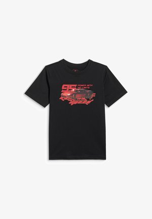 Chłopiec z krótkimi blond włosami, ubrany w czarny T-shirt z grafiką samochodu, czerwone spodnie dresowe, białe skarpety i czarno-białe trampki, stojący na białym tle.